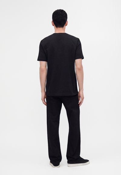 Homme debout de dos, portant un t-shirt noir uni à manches courtes, un pantalon noir et des chaussures noires à semelles blanches sur un fond blanc.