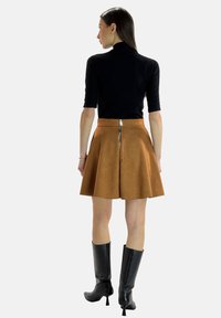 Falda acampanada de ante marrón con un zipper plateado en la parte posterior, combinada con una blusa ajustada negra y botas de tacón negras hasta la rodilla.