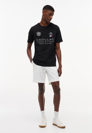 Uomo in piedi che indossa una maglietta nera del team Cadillac Formula 1 Tommy Hilfiger, pantaloncini bianchi, calzini bianchi e scarpe da ginnastica bianche su sfondo bianco.