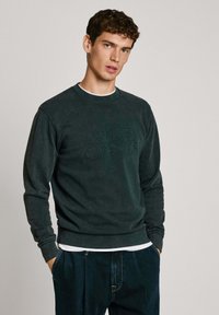 Sudadera verde oscuro hecha de una tela suave, con puños y dobladillo acanalados, logo en relieve "Pepe Jeans" en el frente, superpuesta a una camisa blanca.