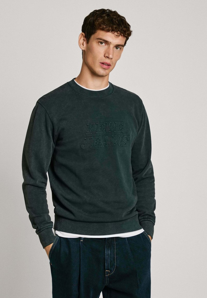 Sudadera verde oscuro hecha de una tela suave, con puños y dobladillo acanalados, logo en relieve "Pepe Jeans" en el frente, superpuesta a una camisa blanca.