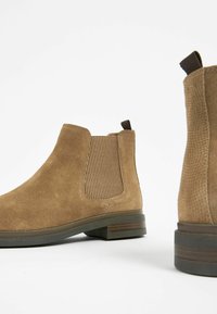 Botas Chelsea de gamuza marrón con paneles laterales estriados, lengüeta y tacón apilado. Acabado texturizado con una suela oscura contrastante.