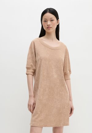 Vrouw met lang zwart haar die een loszittende, beige, korte mouwen badstofjurk draagt tegen een effen witte achtergrond.