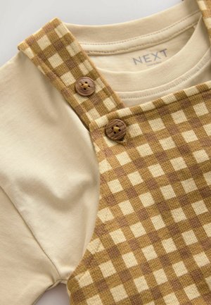 Braun- und cremefarbene kariert Überalls mit Knopfdetails, kombiniert mit einem schlichten cremefarbenen T-Shirt. Glattes Material mit leichter Textur.
