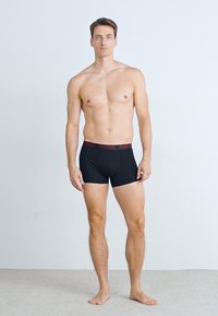 Fekete boxeralsó bordó elasztikus derékpánttal, rajta a márkanevével, férfi modellen viselve, semleges háttér előtt.