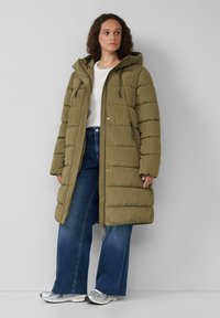 Olivgrüne gepolsterte Parkajacke mit Kapuze, Reißverschluss und Zugband-Details; verfügt über ein gestepptes Design und seitliche Reißverschlüsse. Tragen über blauen Jeans und Turnschuhen.