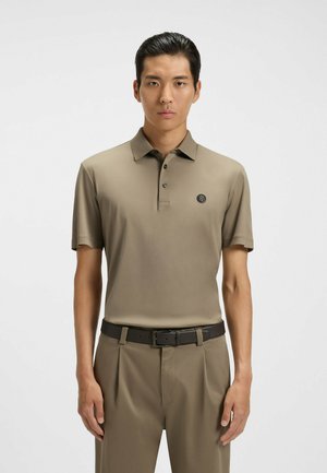 Khakifarbenes Poloshirt mit Kragen, drei Knöpfen und einem schwarzen Emblem. Kombiniert mit beigen Hosen und einem dunkelbraunen Gürtel. Glatte Stoffstruktur.
