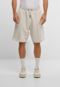 Béžové teplákové šortky s elastickým pásom, sťahovacou šnúrkou, bočnými švami a manžetami na leme, kombinované s bielymi ponožkami a teniskami.