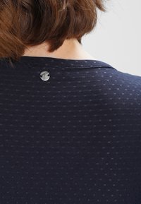 Lyhyet ruskeat hiukset ja vaalea iho näkyvät tummansinisen teksturoituneen kankaan yllä, jossa on pieni pyöreä musta ja hopeinen nappi kauluksen lähellä.