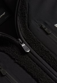 Zwarte fleece-voering jas met gladde stof, meerdere ritszakken en geprofileerde accenten. Label geeft maat medium aan.