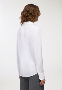 Camicia bianca a maniche lunghe con una texture liscia, caratterizzata da un colletto classico, polsini con bottoni e un design con spallina sul retro.