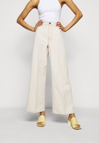 Light beige wide-leg jeans made from denim, featuring a high waist, front pockets, and a subtle fade. Paired with yellow open-toe heels.