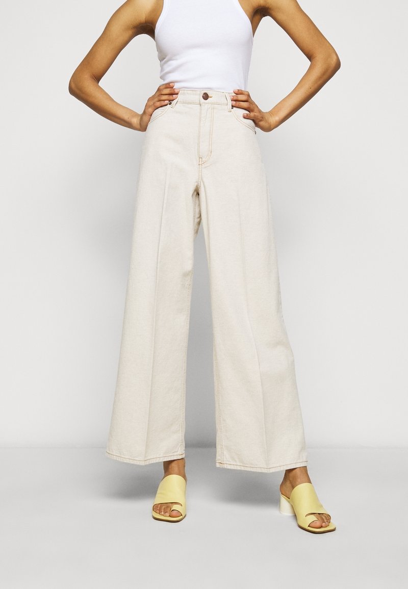 Light beige wide-leg jeans made from denim, featuring a high waist, front pockets, and a subtle fade. Paired with yellow open-toe heels.