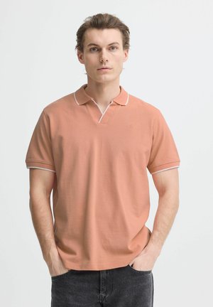 Man with light skin and light brown hair wears salmon-colored short-sleeve polo shirt and dark jeans, standing with hands in pockets.