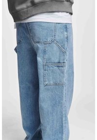 Pantalones cargo de mezclilla azul claro con múltiples bolsillos, corte relajado y detalles de costura visibles. La tela parece texturizada y resistente.
