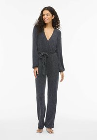 Juodas metalinis blizgučiais puoštas kombinezonas su ilgomis rankovėmis, gilia V formos iškirpte, juosiamas diržu juosmuo ir plačiomis kelnėmis. Lygios tekstūros, aptemptas siluetas.