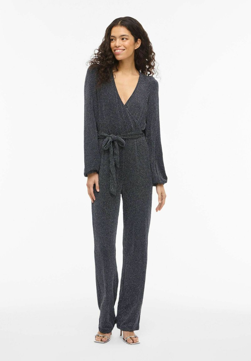 Juodas metalinis blizgučiais puoštas kombinezonas su ilgomis rankovėmis, gilia V formos iškirpte, juosiamas diržu juosmuo ir plačiomis kelnėmis. Lygios tekstūros, aptemptas siluetas.