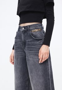Camiseta negra corta combinada con jeans gris de tiro alto y perneras anchas, que cuentan con un detalle de cadena dorada en el bolsillo frontal. Textura de denim suave.