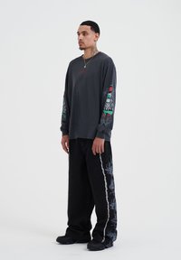Graues Langarmshirt mit bunten Grafikdrucks auf den Ärmeln, kombiniert mit lockeren schwarzen Hosen mit dezentem weißen Muster und schwarzen Schuhen.