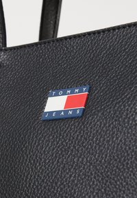Schwarze, strukturierte Lederhandtasche mit einem rechteckigen Logo-Patch in Blau, Rot und Weiß, das mit "TOMMY JEANS" beschriftet ist.