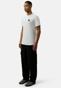 Weißes Baumwoll-T-Shirt mit kurzen Ärmeln, das auf der linken Brust eine kleine schwarz-goldene Logo-Patch aufweist. Kombiniert mit lockeren schwarzen Cargo-Hosen.