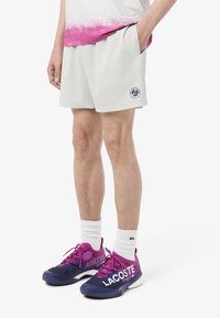 Pantalones cortos deportivos gris claro con un patrón texturizado y un parche de logo. Combinados con zapatillas moradas y marinas con logotipos blancos y calcetines blancos.