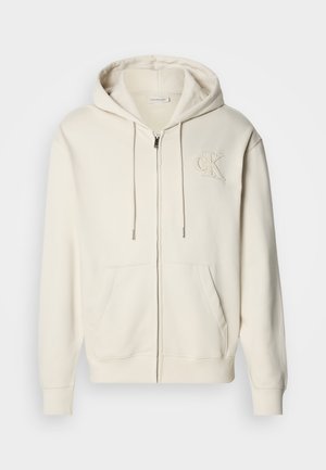 Krémově zbarvená mikina na zapínání na zip s kapucí na stažení šňůrkou, předními kapsami a vyšívaným logem na hrudi. Měkká látka a volný střih.