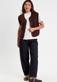 Young woman wearing a brown fleece vest over a white t-shirt, dark wide-leg jeans, and brown sneakers against a plain white background.