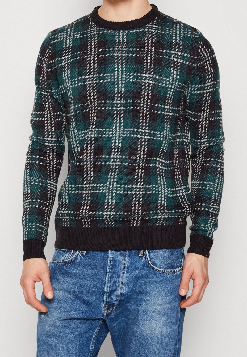 Sweter dziany w zieloną, czarno-białą kratę, z okrągłym dekoltem oraz ściągaczami na rękawach i u dołu. Noszony razem z niebieskimi dżinsami.