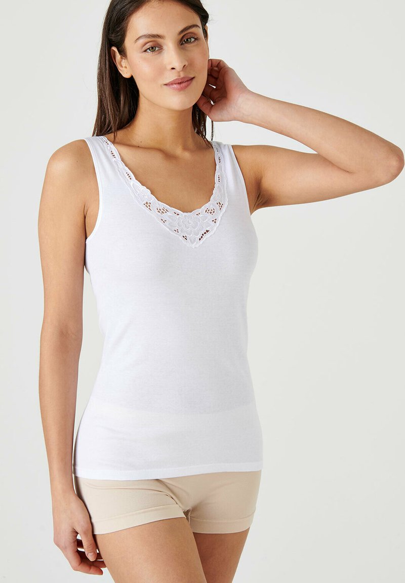 Top bianco senza maniche con scollatura a V e rifiniture in pizzo, caratterizzato da motivi floreali. Il tessuto è liscio e aderente, abbinato a pantaloni corti beige.