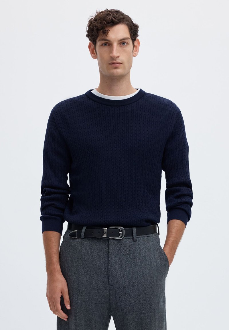 Homme aux cheveux bouclés portant un pull à torsades bleu marine, un pantalon gris et une ceinture noire, debout une main dans la poche.