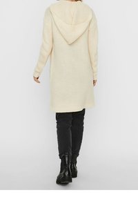 Długi, kremowy sweter z kapturem, wykonany z dzianiny o fakturowanym splocie z prążkowanym wykończeniem na dole, noszony na ciemnych spodniach i czarnych botkach za kostkę.