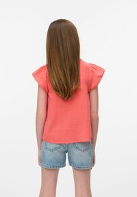 Top corto corallo con maniche a sbuffo, realizzato in tessuto testurizzato. Abbinato a pantaloni corti di jeans chiari. La vista posteriore mostra l'orlo della maglietta.