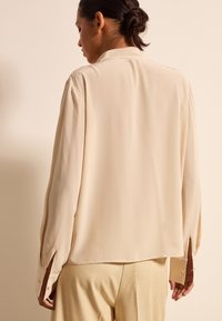 Beige Seidenbluse mit langen Ärmeln, geschlitzten Manschetten und lockerer Passform. Sanfte Textur, minimale Details und weicher Fall.