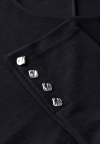 Tessuto a maglia blu navy con superficie testurizzata e quattro bottoni argentati lucidi lungo il lato. Scollo rotondo semplice con dettagli a costina.