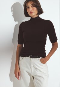 Woman with short brown hair wearing a dark brown turtleneck and white pants, standing with one hand in pocket against a white wall.
