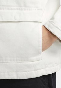 Chaqueta de denim blanca con un bolsillo en el pecho, corte relajado y detalle de puños doblados. La tela parece resistente con una textura suave.