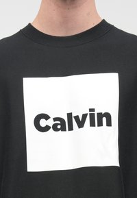 Čierne tričko s okrúhlym výstrihom, ktoré má veľkú bielu obdĺžnikovú grafiku s nápisom "Calvin" v tučných čiernych písmenách. Mäkká textúra, ležérny dizajn.