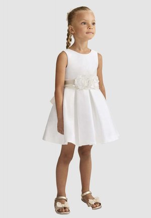 Giovane ragazza con capelli intrecciati che indossa un vestito bianco senza maniche con cintura floreale e sandali dorati, in piedi davanti a uno sfondo semplice.