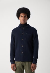 Mann mit Afro-Haarschnitt, der eine navyblaue strukturierte Knopfvorderjacke über einem schwarzen Rollkragenpullover und olivgrünen Hosen trägt und vor einem schlichten Hintergrund steht.