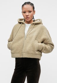 Beige, textured fleece hoodie with a cropped design, large hood, and front pockets. Features a zip closure and drawstring accents.