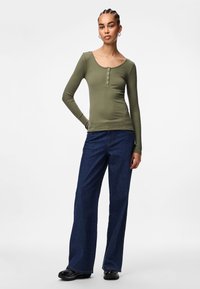 Olivgrünes Longsleeve-Shirt mit figurbetonter Passform, gerippter Textur und Knopfdetails, kombiniert mit weit geschnittenen, dunkelblauen Jeans. Schwarze Schuhe runden das Outfit ab.