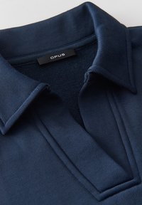 Col et encolure de polo bleu marine avec une étiquette noire portant la mention "OPUS" sur fond de tissu texturé.