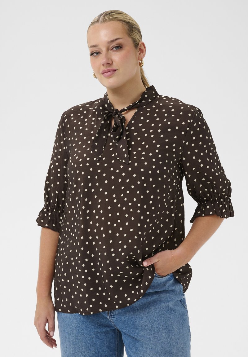 Femme aux cheveux blonds portés en queue de cheval, vêtue d'un chemisier noir à pois blancs avec un col noué, associée à un jean bleu, debout avec une main dans la poche.