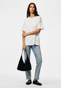 Woman with curly hair wearing a white oversized t-shirt, light blue jeans, black shoes, and holding a black shoulder bag.