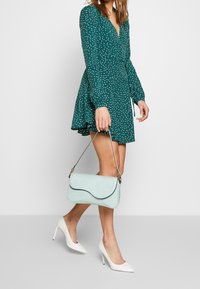 Vestido verde con lunares blancos, mangas largas y dobladillo acampanado; sosteniendo un bolso de mano de cocodrilo en un tono menta claro; usando tacones altos blancos.