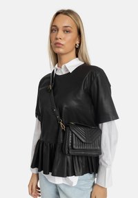Schwarzes Kunstleder-Peplum-Oberteil mit kurzen Ärmeln über einem weißen Kragenhemd. Das Model trägt eine kleine schwarze gesteppten Crossbody-Tasche.