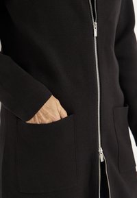 Svart zip-front kofta i texturerad tyg, med sidofickor och en str�ömlinjeformad siluett. Silvrig dragkedja framhäver designen.