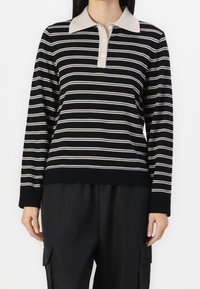 Person som bär en svart och vit randig långärmad polo skjorta med beige krage och svarta cargobyxor, stående mot en vit bakgrund.