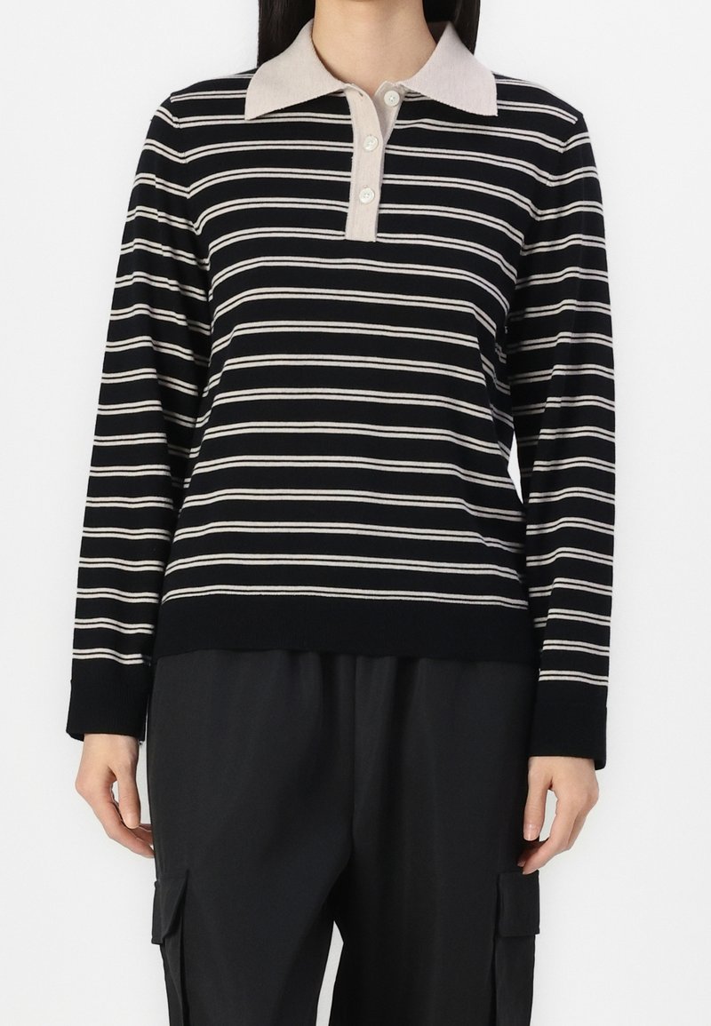 Person som bär en svart och vit randig långärmad polo skjorta med beige krage och svarta cargobyxor, stående mot en vit bakgrund.